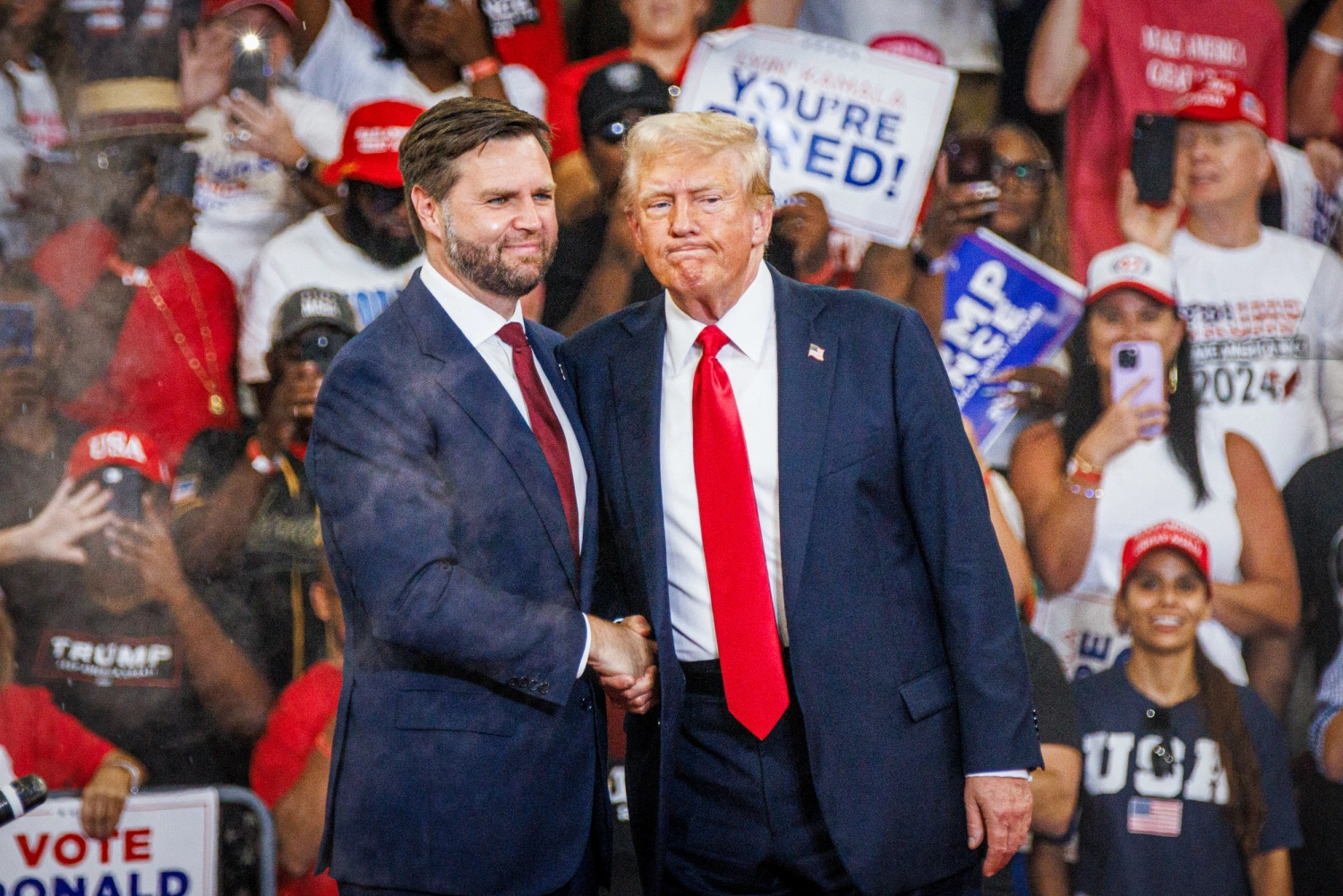 Former President Donald Trump's Vance 2024 campaign. Trump, in a dark suit and red tie, stands beside Vance, who is wearing a dark suit and light blue tie. They are smiling and shaking hands.